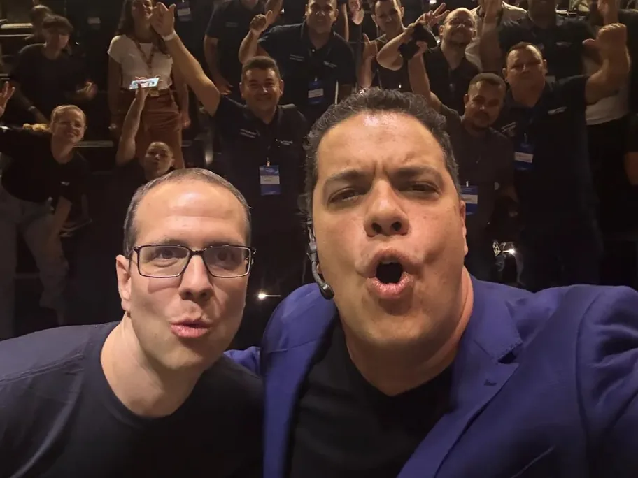 Selfie de dois palestrantes com o time do Mercado Livre comemorando ao fundo
