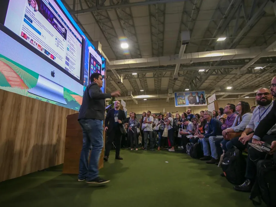 Palestrante falando para o público em um pavilhão de evento da Salesforce, com telões ao fundo